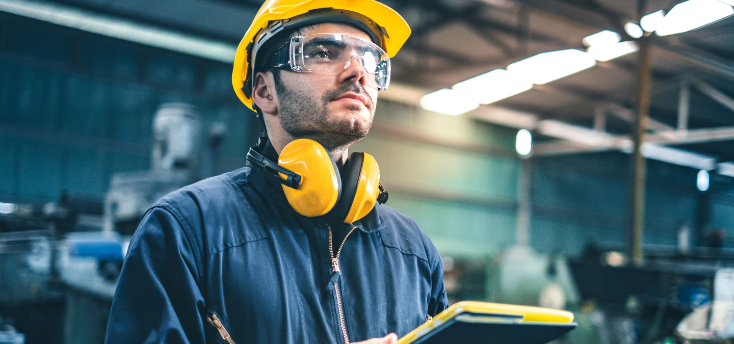 Opérateur en industrie équipé de protections auditives et visuelles, tenant une tablette dans un atelier de production
